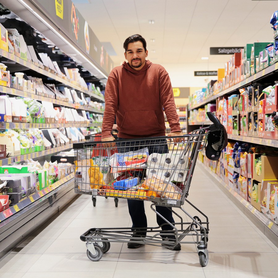 Raphael steht in einem Supermarkt-Gang, er stützt sich auf einem vollen Einkaufswagen ab