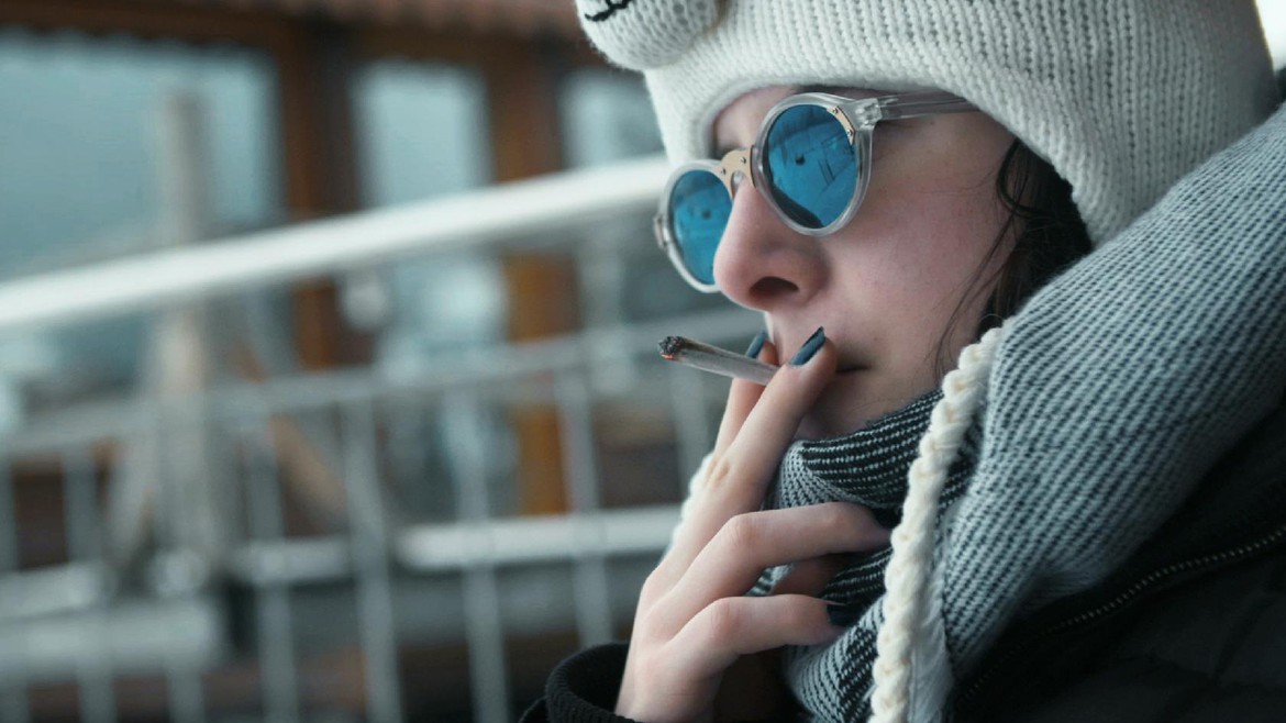 Magda-Lena sitzt auf der Terrasse und raucht. Sie trägt eine dicke Jacke Kappe und Sonnenbrille.