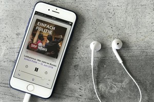 Mobiltelefon mit Podcast-App auf der «Einfach Politik» läuft, daneben Kopfhörer