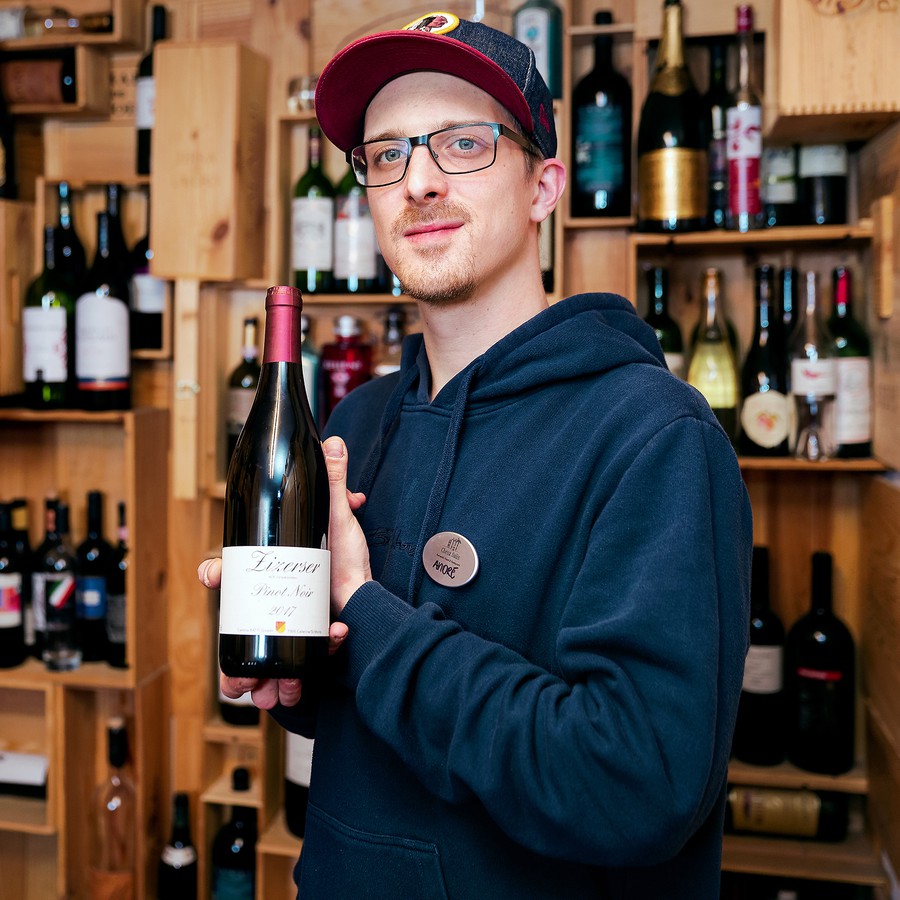 André steht vor einem grossflächigen Weinregal und hält eine Flasche Rotwein in die Kamera