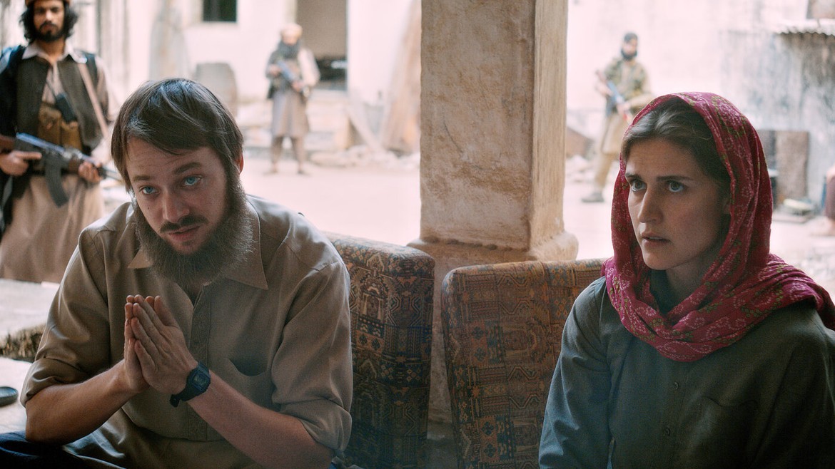 David und Daniela sitzen auf Sesseln, hinter ihnen läuft ein bewaffneter Taliban durch