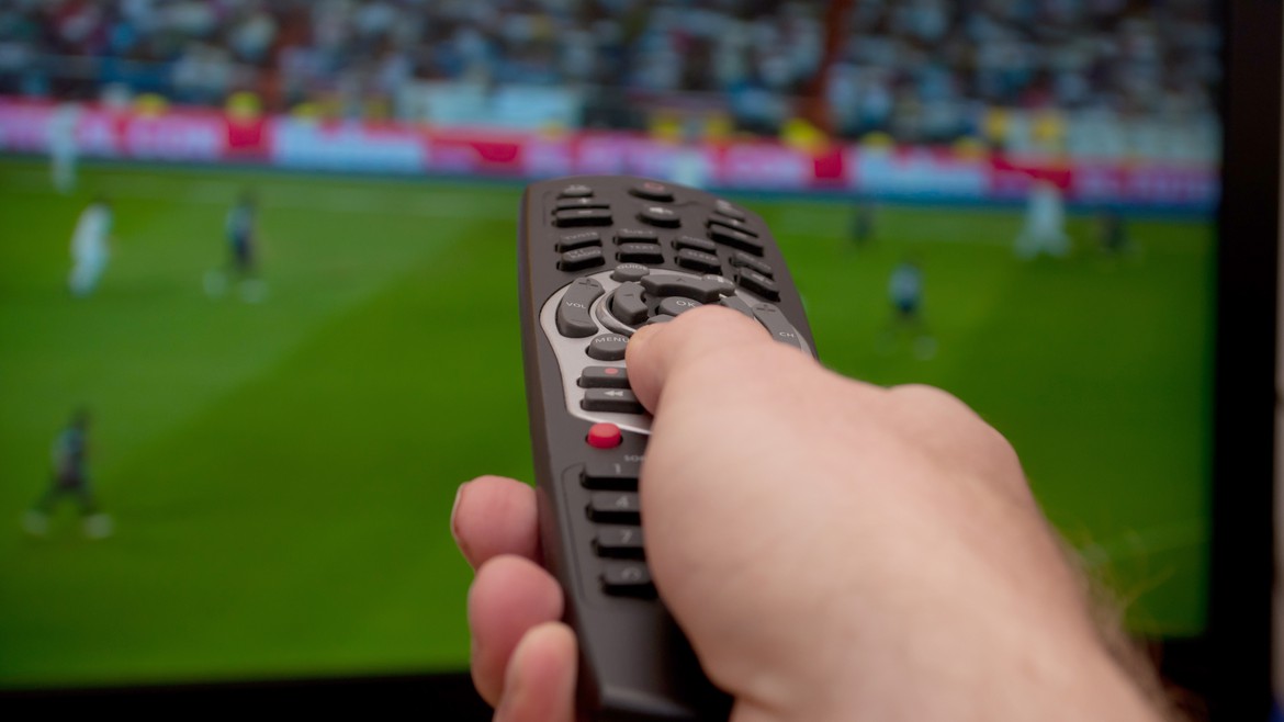 Fernbedienung zeigt auf Fernseher, auf welchem verschwommen ein Fussballspiel zu sehen ist.