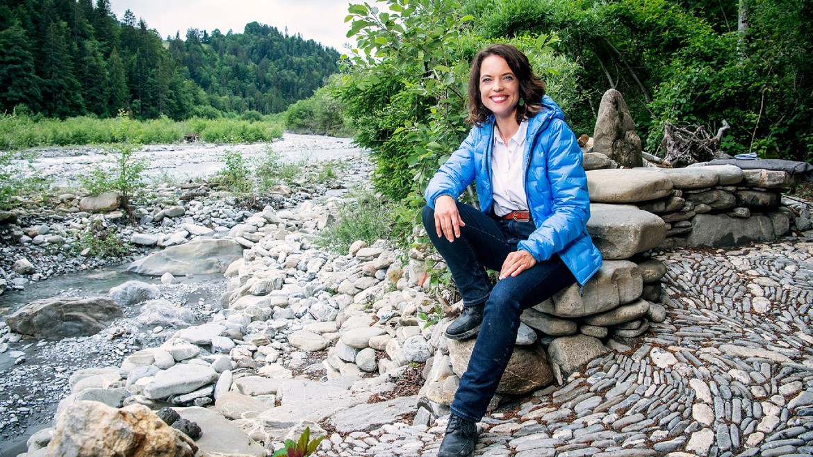Mona Vetsch sitzt auf einem Steinhaufen an einem Fluss und lächelt in die Kamera