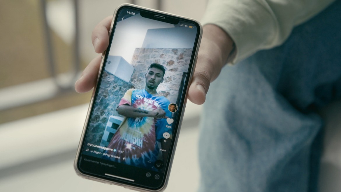 Ein Handy wird ins Bild gehalten mit einem laufenden Video von Younes Saggara.