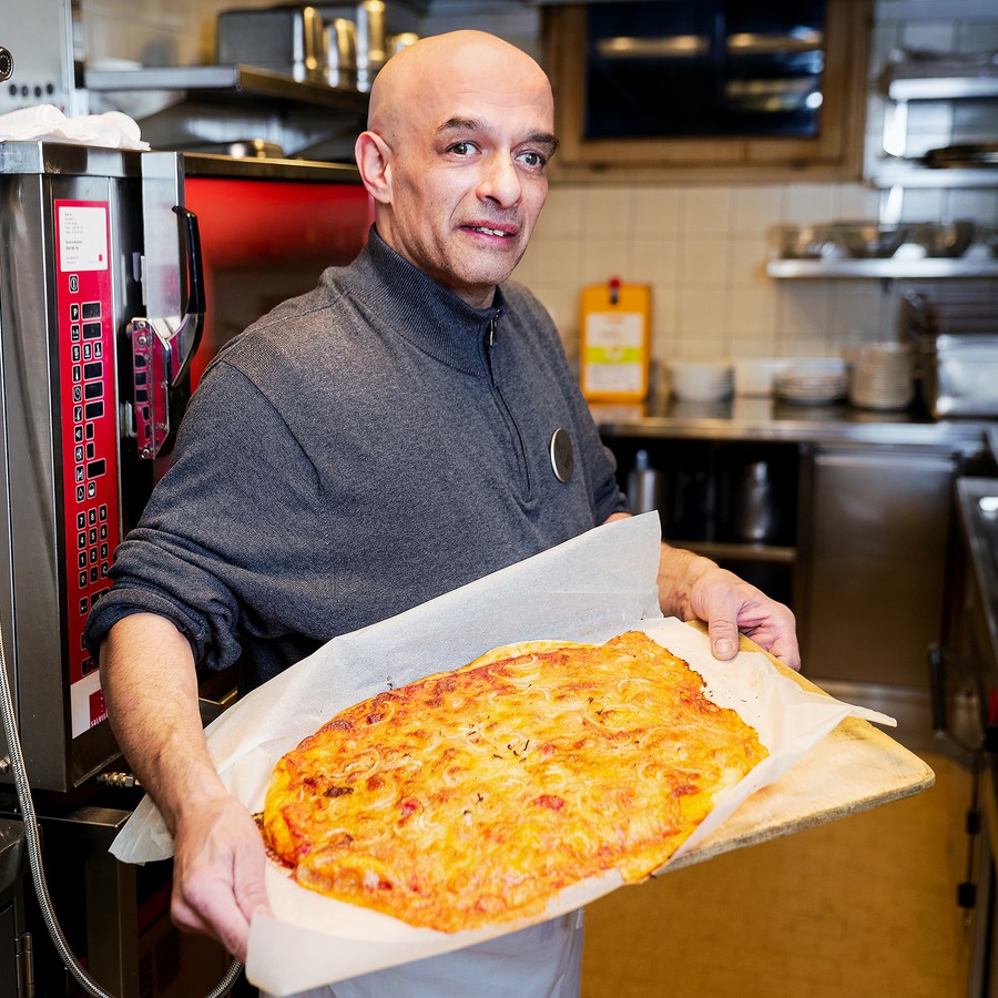 Ilker steht in der Hotelküche und hält ein grosses Blech mit einer gebackenen Pizza in den Händen