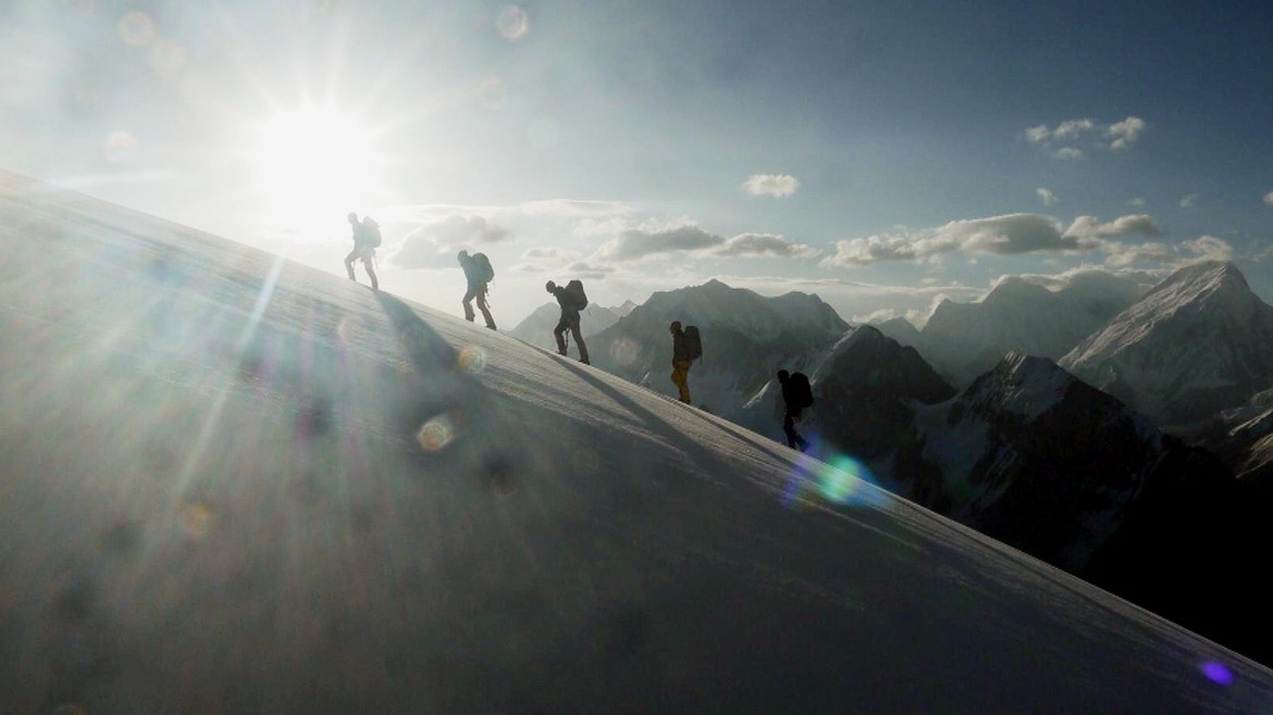 Fünf Bergsteiger gehen Berghang hinauf, die Sonne scheint in die Kamera