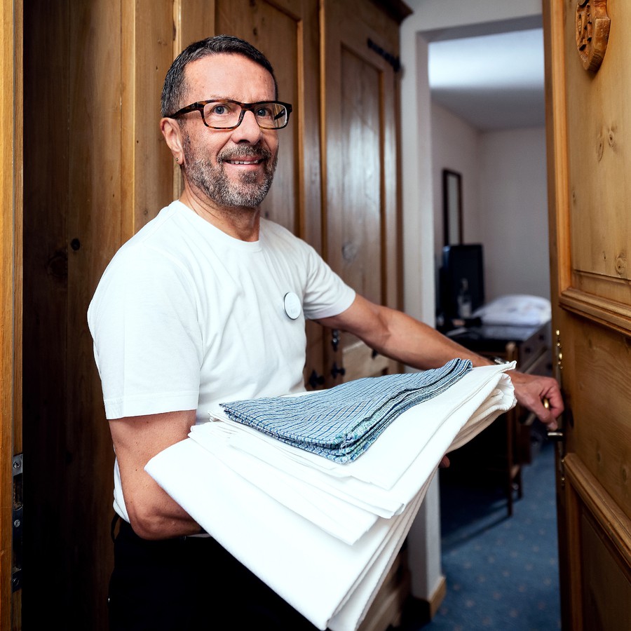 Ruedi steht lächelnd vor der geöffneten Zimmertür und hat einen Stapel frisch gebügelter Laken auf dem Arm