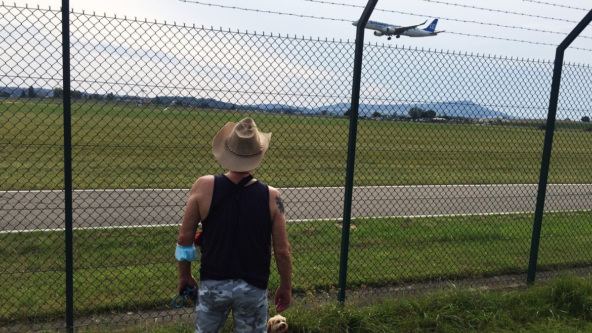 Edi steht vor einem Zaun und beobachtet ein vorbeifliegendes Flugzeug