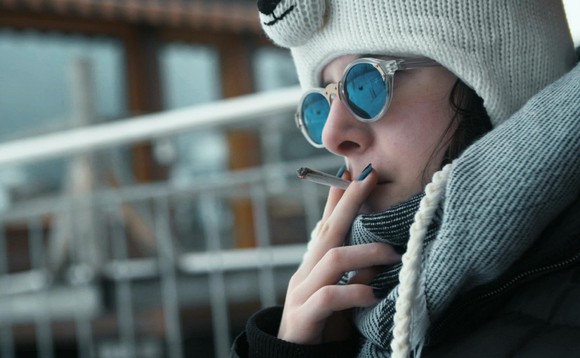 Magda-Lena sitzt auf der Terrasse und raucht. Sie trägt eine dicke Jacke Kappe und Sonnenbrille.